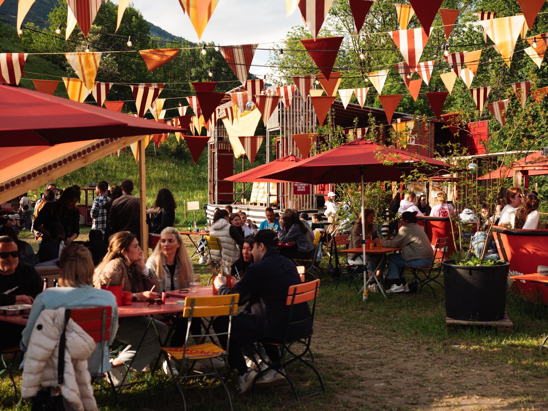 Outdoor café with people seated under red umbrellas and colorful bunting in a sunny garden setting