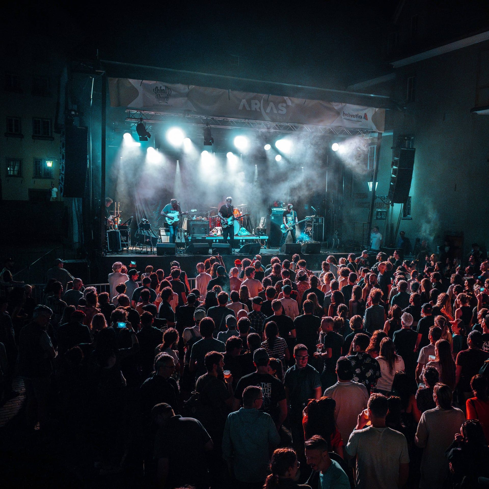 Ein dicht gedrängtes Konzertpublikum steht vor einer hell erleuchteten Bühne mit einer Live-Band unter blau-weißem Licht.