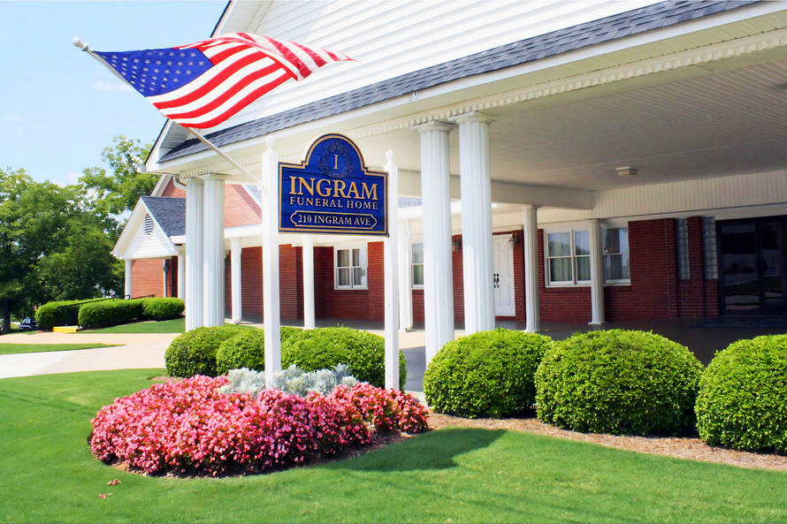 Ingram building entrance with American flag, white columns, brick facade, and landscaped shrubs