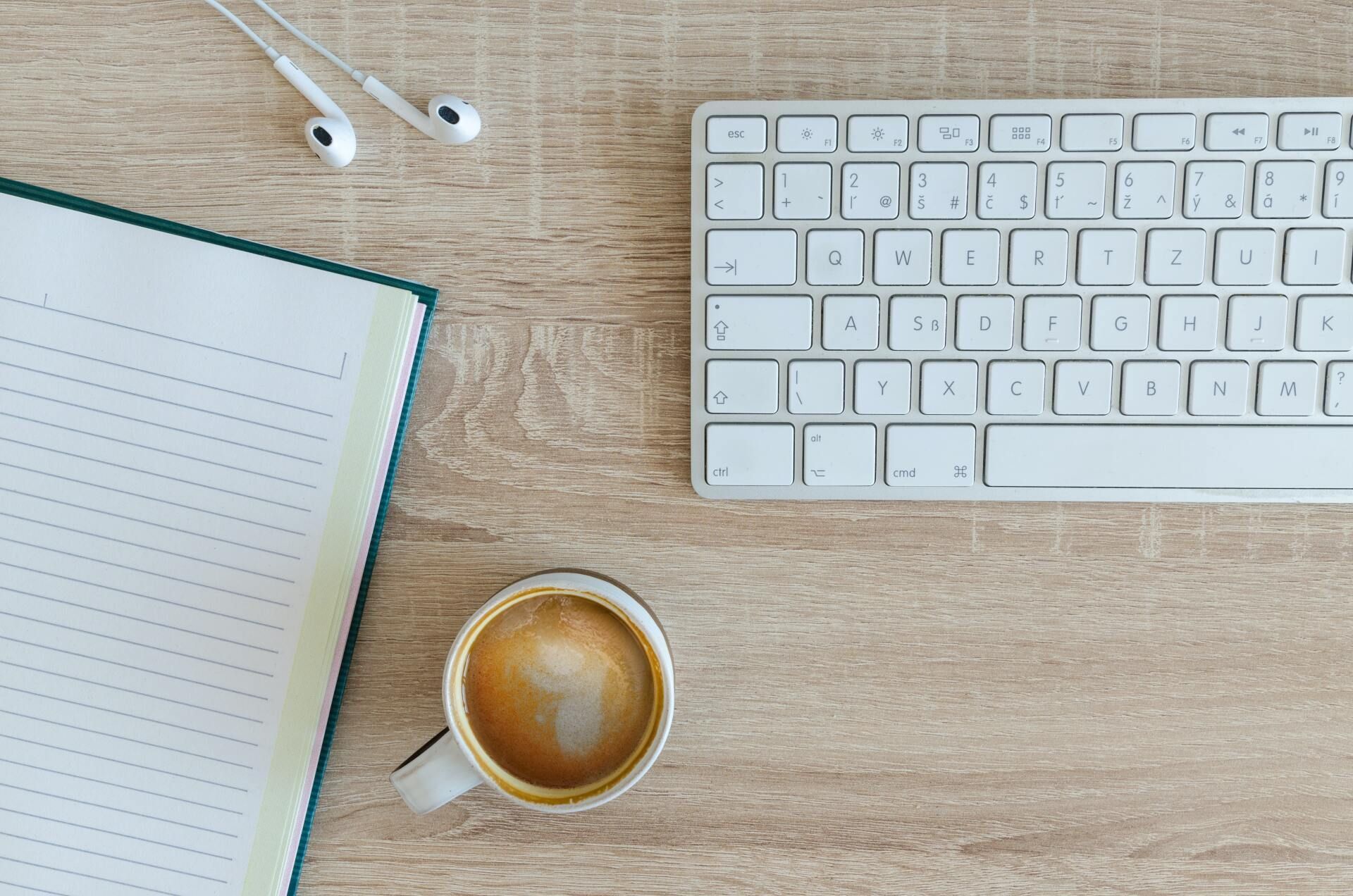 Top-down desk with notebook, coffee cup, white keyboard, and earbuds on a light wood surface.