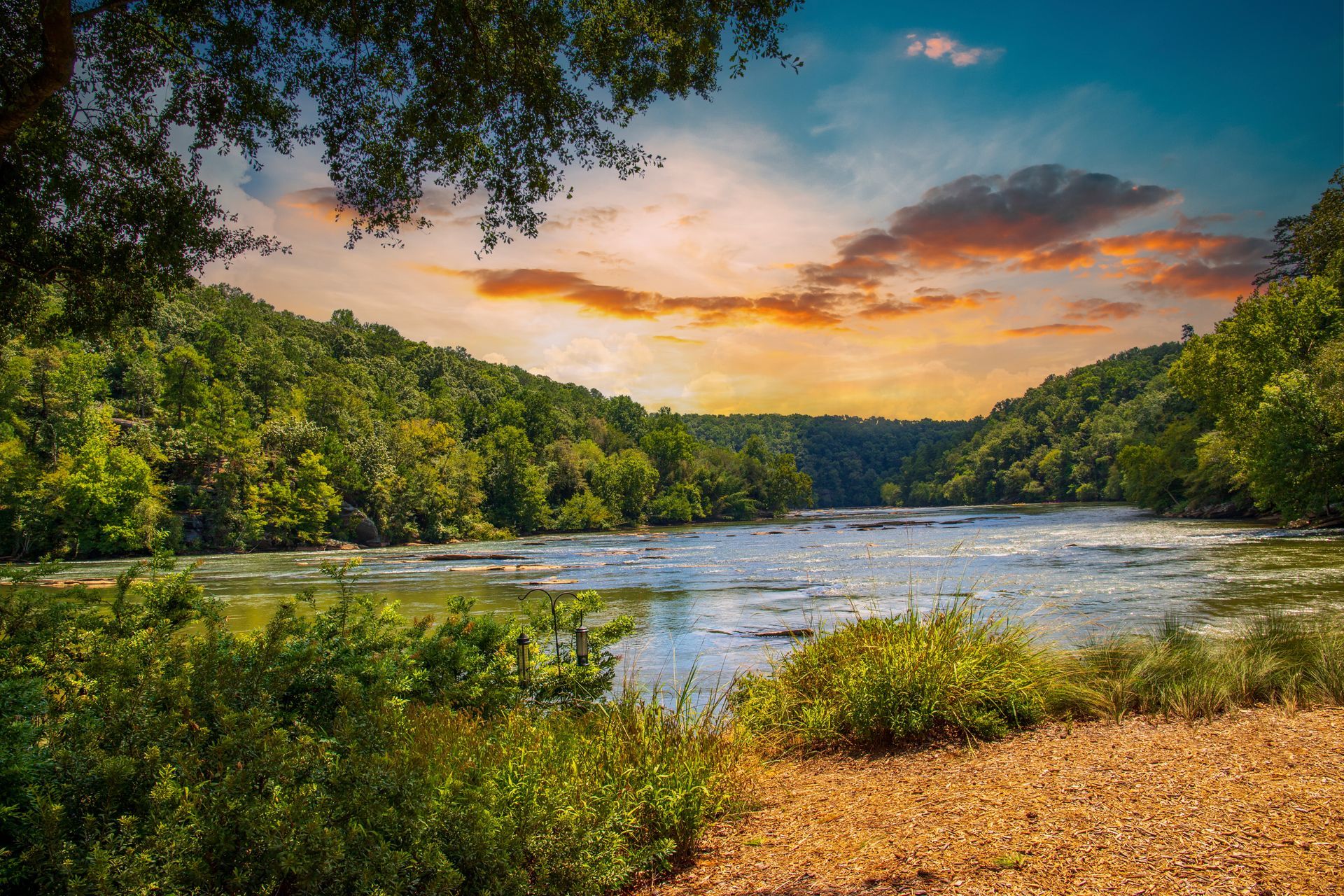 Peaceful river at sunset, with green trees, a sandy bank, and orange clouds overhead