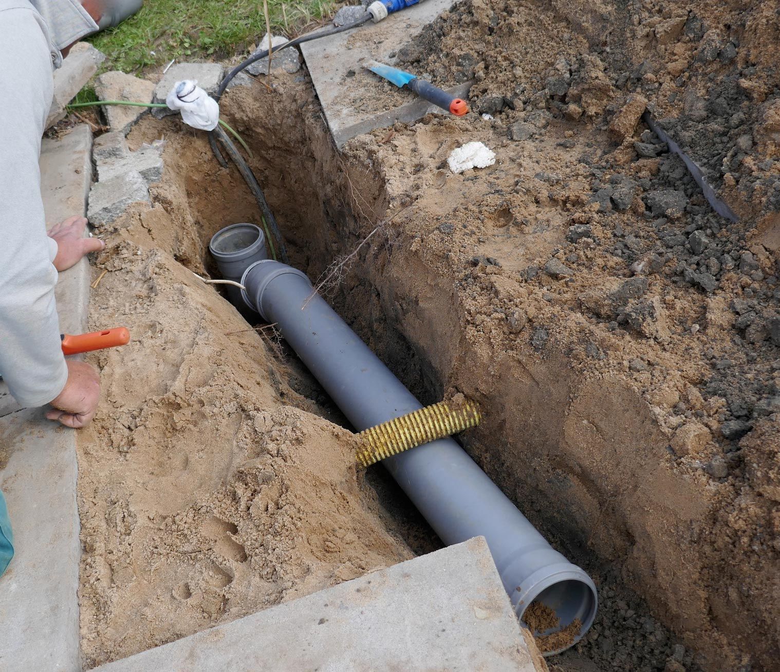 A person installing a gray pipe in a trench filled with dirt.  The pipe is held by a yellow mesh.