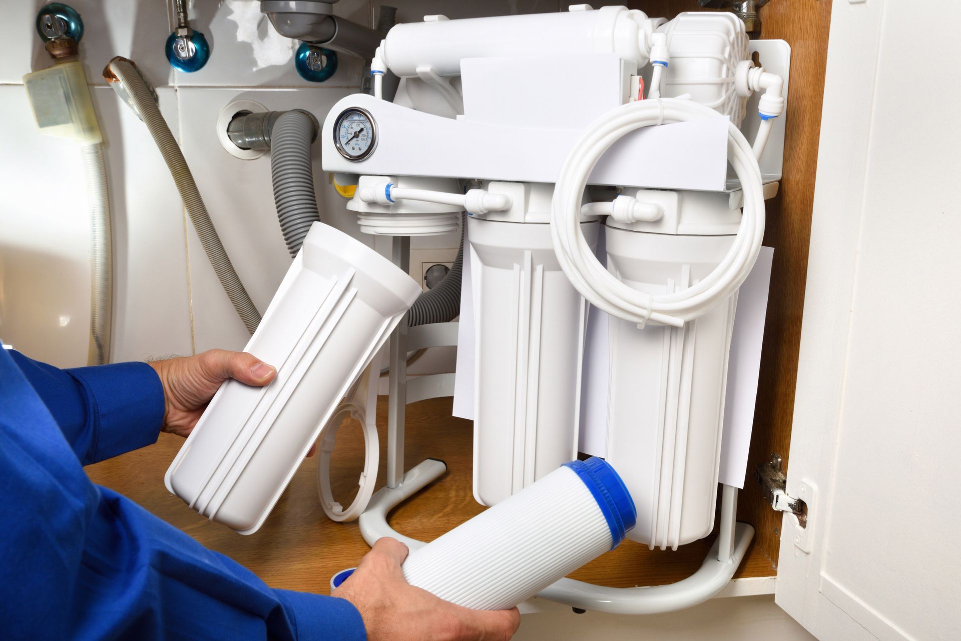 Person replacing water filter cartridges under a sink; white filters and plumbing visible.