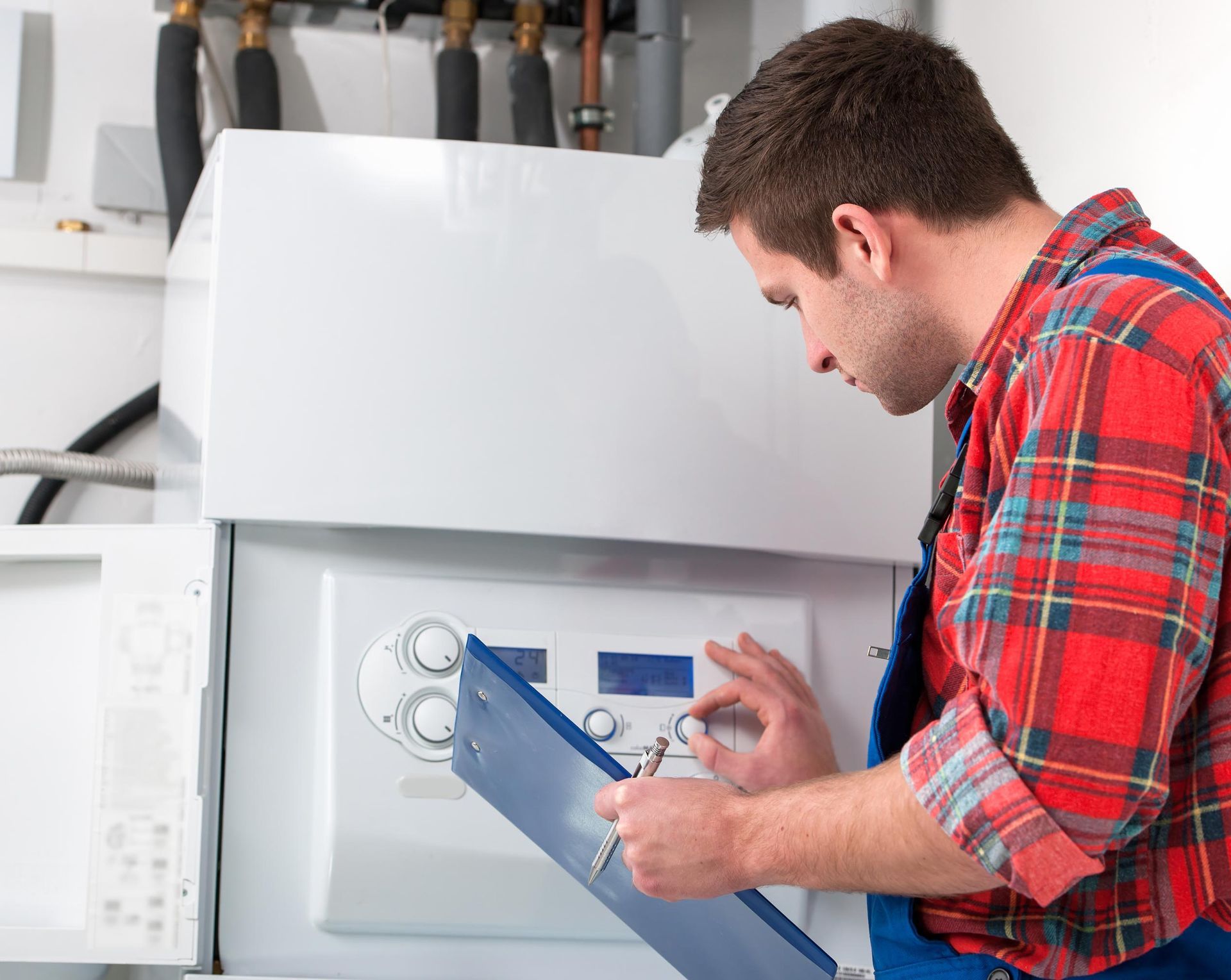 Man in red plaid shirt and blue overalls inspecting a white boiler, holding a clipboard.