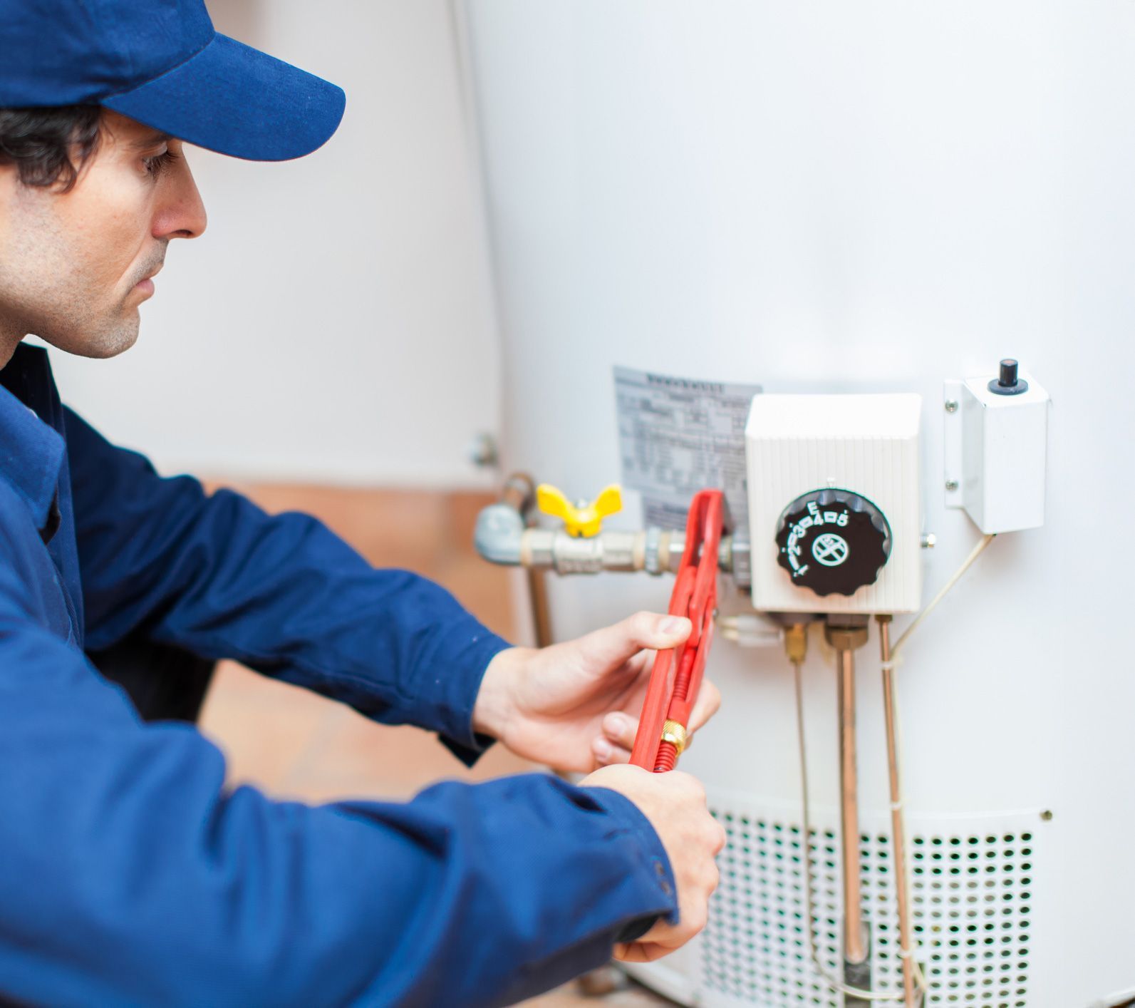 A plumber in blue overalls uses a wrench on a water heater.