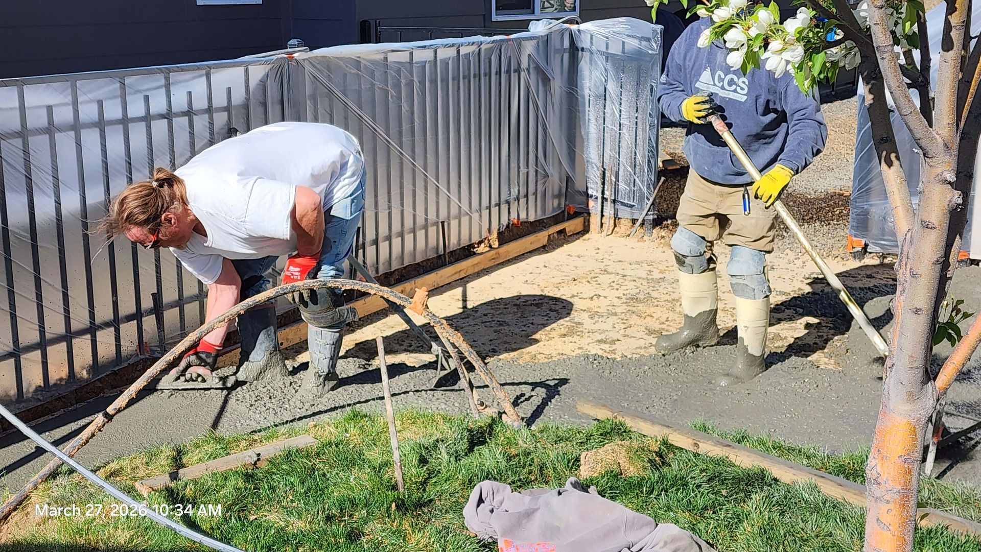 Two people working with tools beside a fenced garden and soil on a sunny day