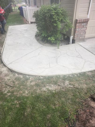Concrete walkway around a shrub beside a house, with a grassy yard in the foreground.