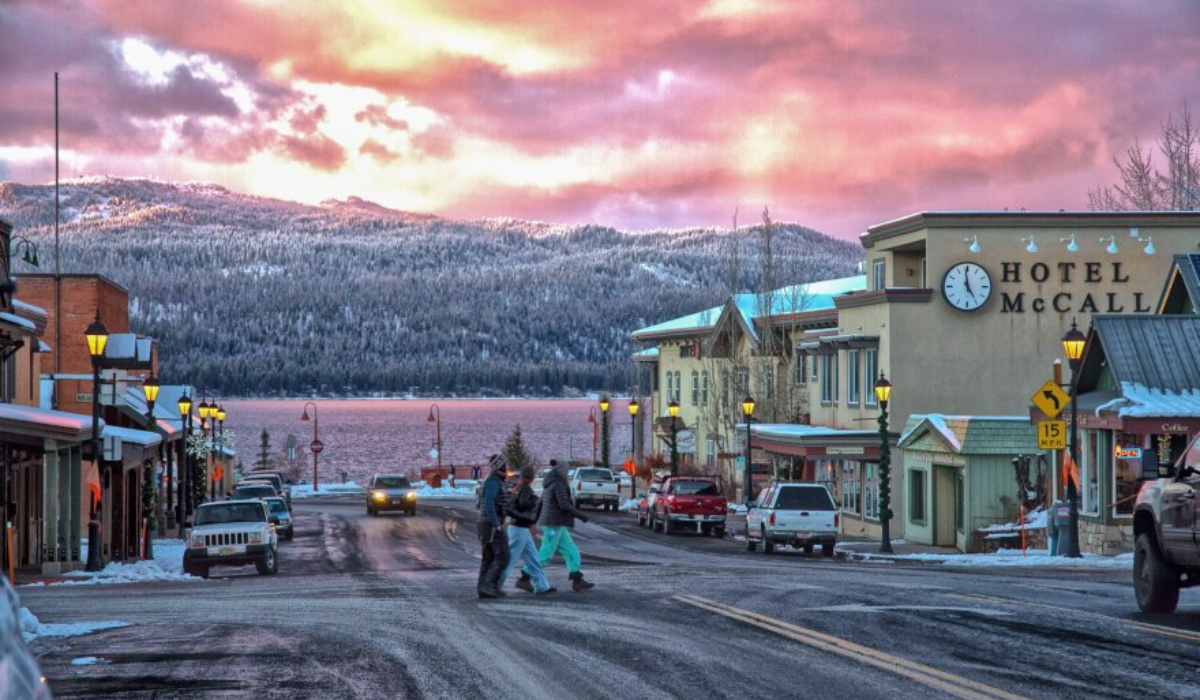 Snowy main street with shops, parked cars, and a hotel under a pink sunset sky