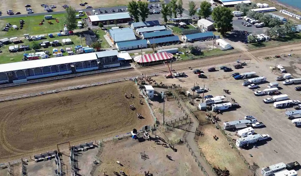 Aerial view of an outdoor fairground with a dirt arena, parking lots, and long white-roofed buildings.