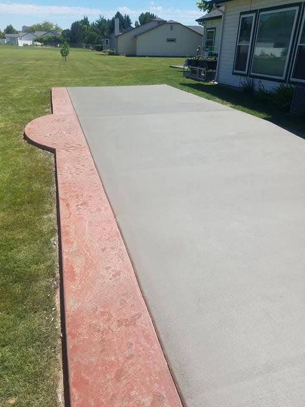 Concrete patio beside a lawn, with a red brick border near a house and a grassy yard in the background