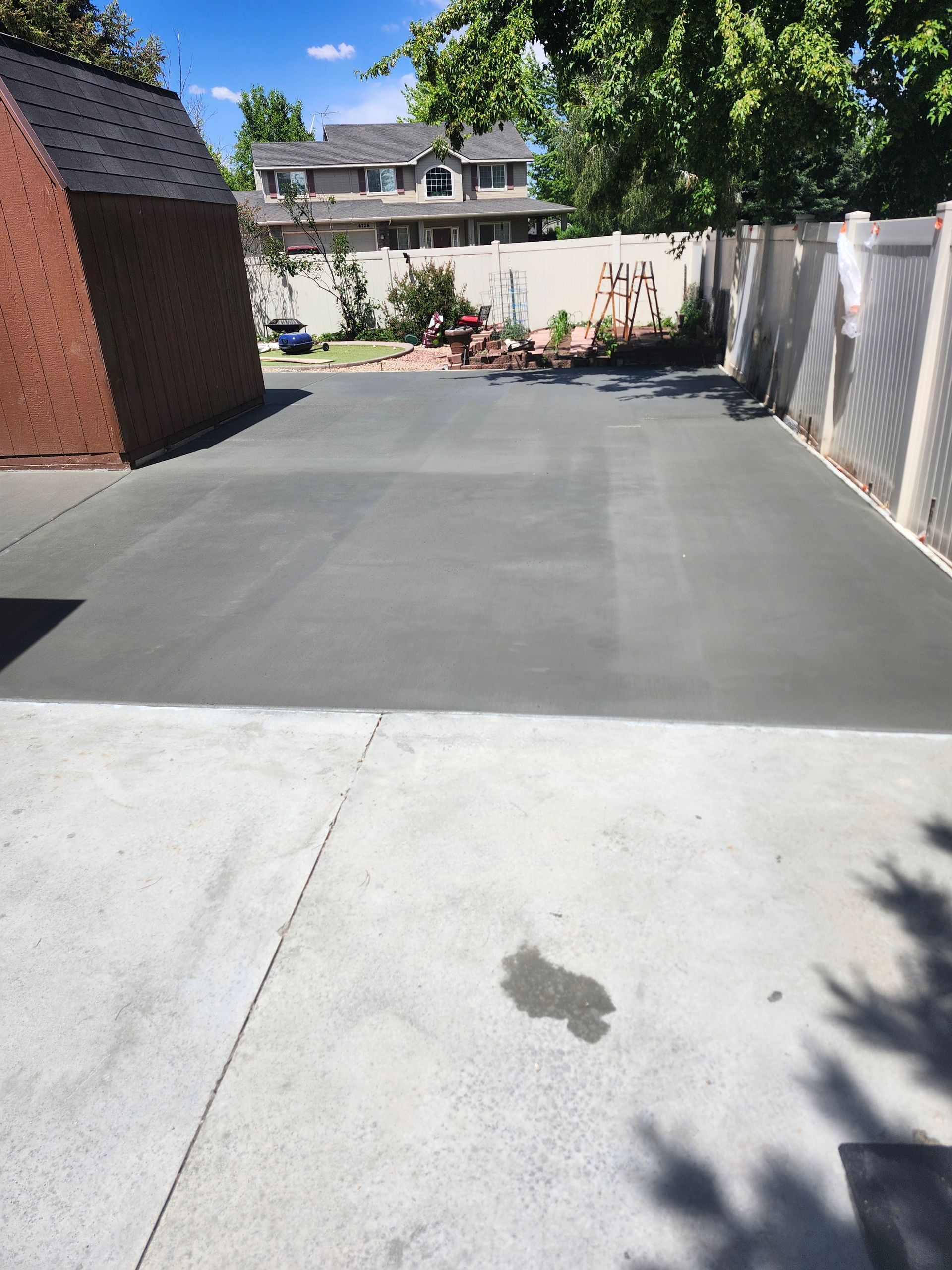 Backyard patio with a large gray concrete slab, white fence, and flower bed in the distance
