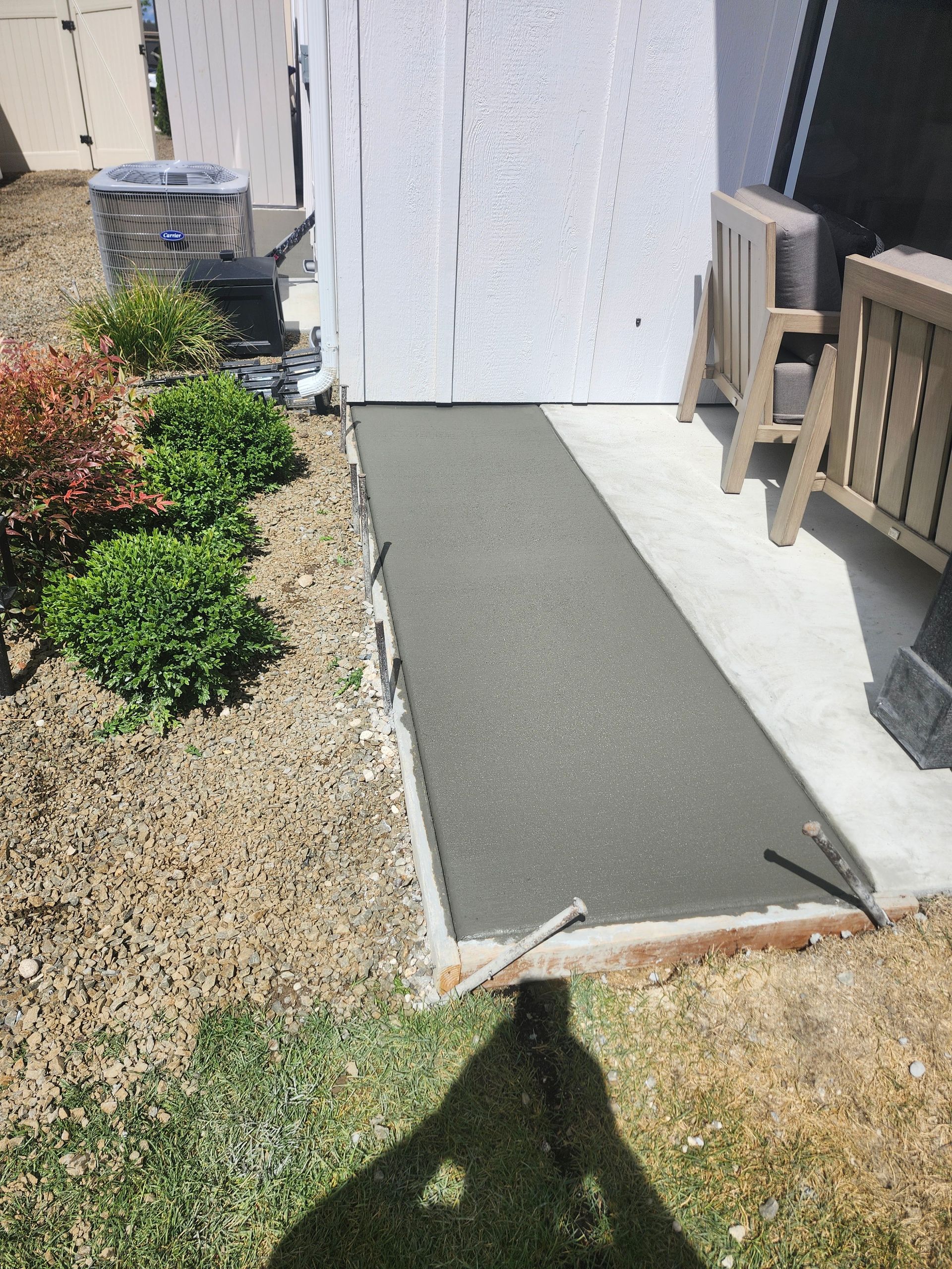 Concrete walkway beside a white house, with a gravel garden bed and shrubs on the left.
