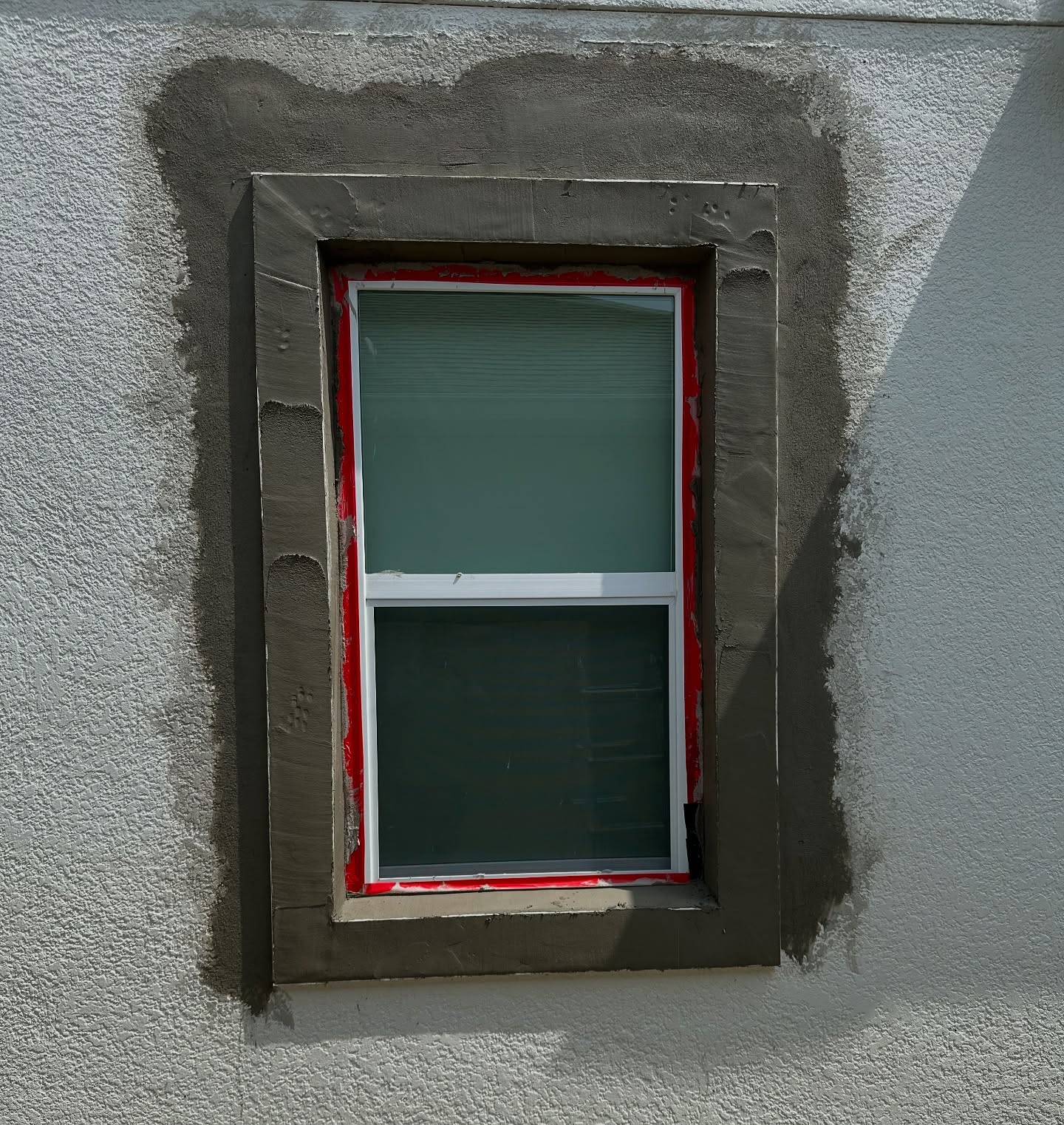 A rectangular window in a textured stucco wall, framed by a layer of wet, dark-grey mortar surrounding the window frame.