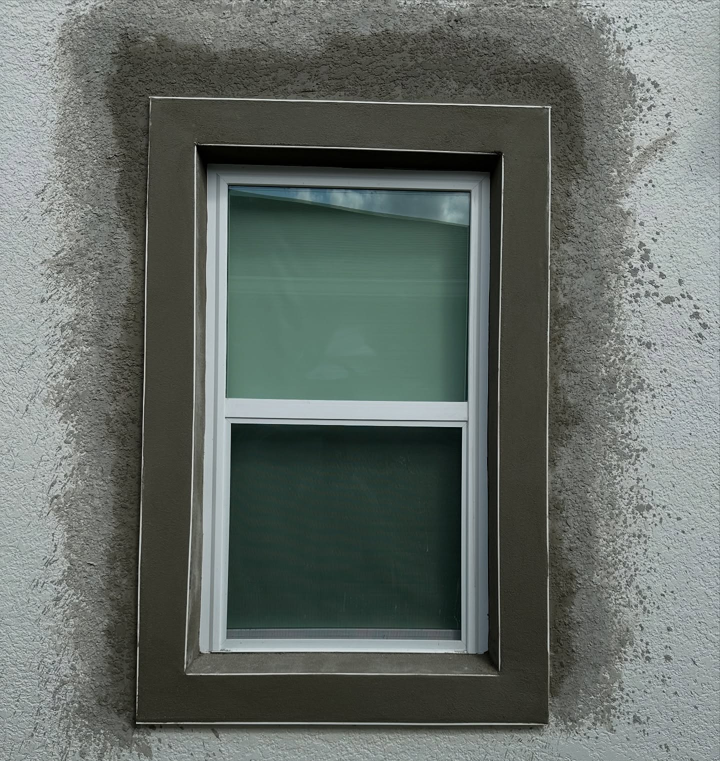 A vertical window with a dark brown frame, set into a wall with rough, light gray stucco plastering around the edges.