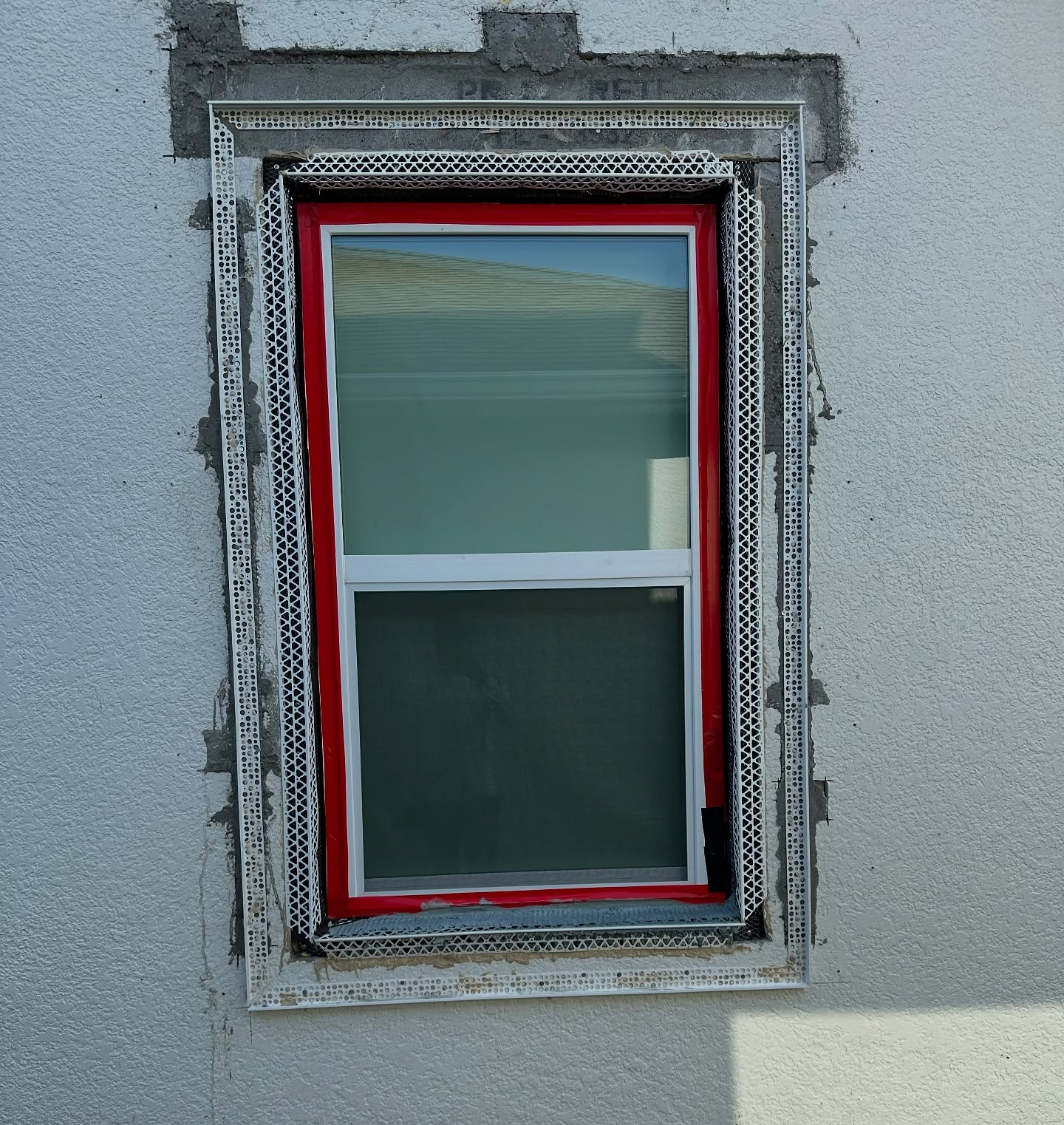 A rectangular window installed in a light gray stucco wall, framed with metal corner bead and red flashing tape.