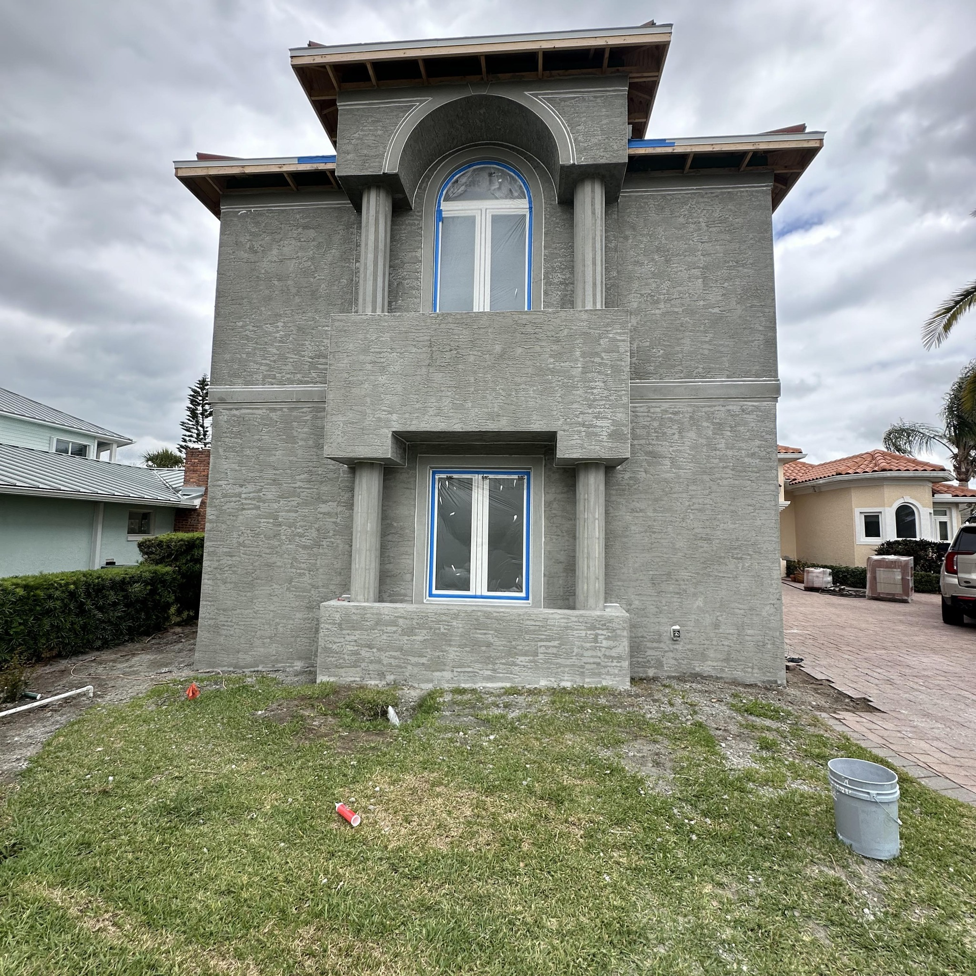 A two-story house under construction with a gray stucco exterior, arched top window, and pillars on a residential street.