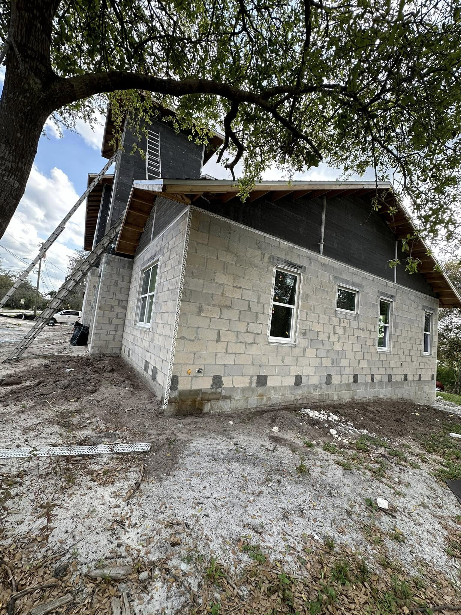 A stone house under construction with a dark gable, partially shaded by a large tree on a cloudy day.