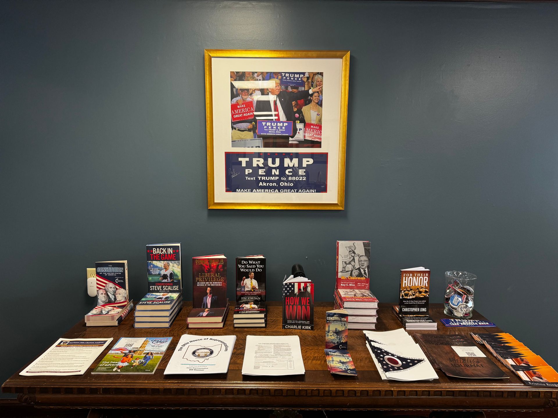Display of Trump memorabilia on a table beneath a framed rally poster, against a blue wall.