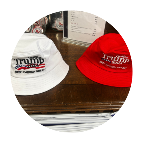 Two Trump 2024 bucket hats on a wooden surface: one white, one red. Both have the slogan, “Keep America Great!”