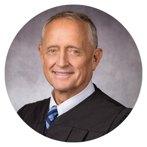 Judge in black robe, white shirt, and blue tie smiling in a formal portrait.