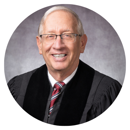 Judge in black robes smiling at the camera, wearing glasses and a tie.