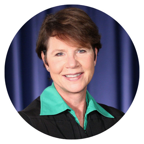 Woman judge with short brown hair smiles, wearing a black robe and green collar, against a blue curtain backdrop.