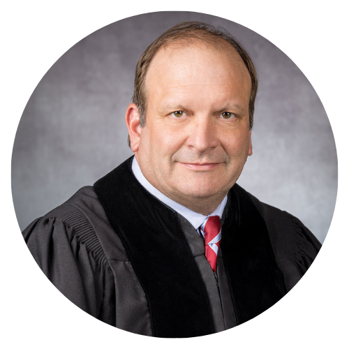 Judge in black robe, red tie, seated, looking forward with a neutral expression.