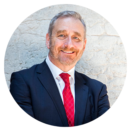 Man in suit and red tie smiling against a stone wall.
