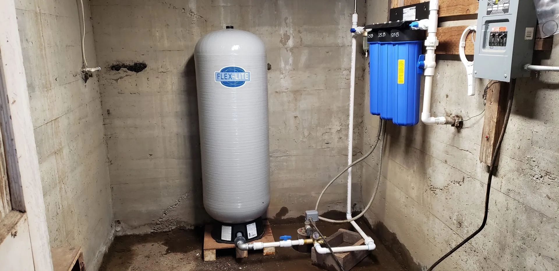 Water filtration system and pressure tank in a concrete room. Blue filter, gray tank, and white pipes are visible.