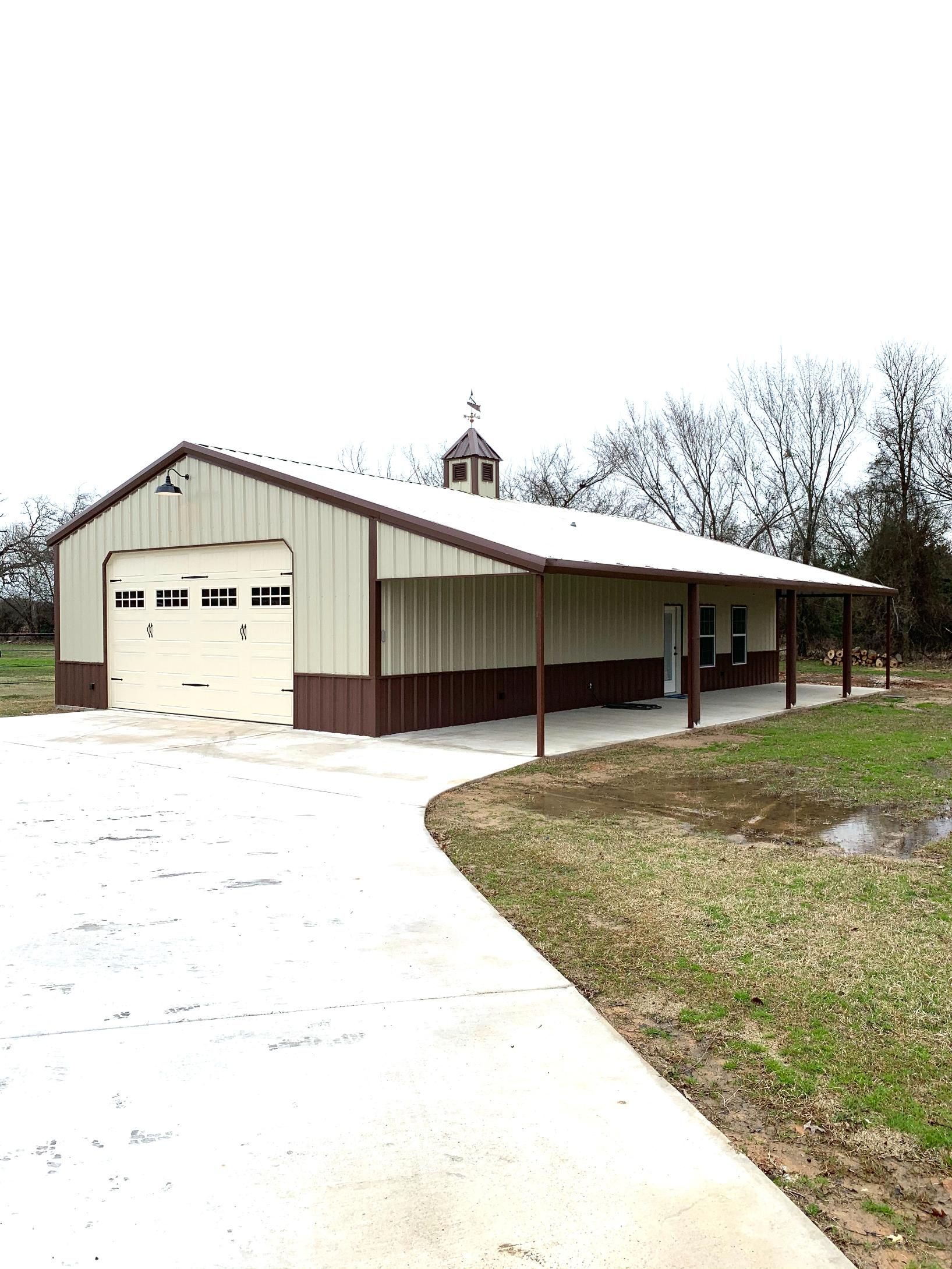 A light-colored metal barn with a brown lower trim, a garage door, and a side porch, set near a concrete driveway.