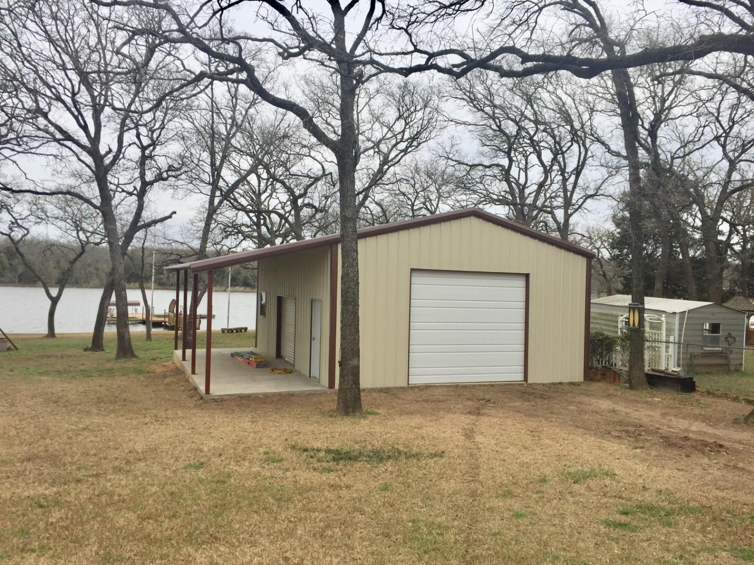 A tan metal workshop with a garage door and side porch, located on a grassy lot near a lake with bare trees.