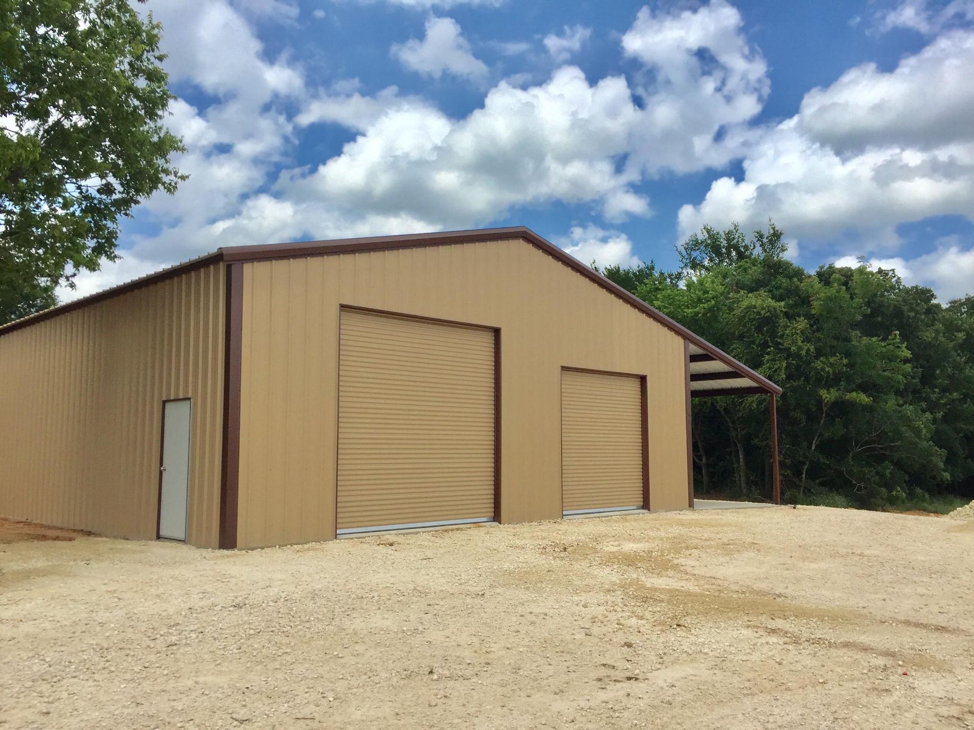 A tan, metal-sided storage building with two roll-up doors and one entry door sits on a gravel lot under a blue sky.