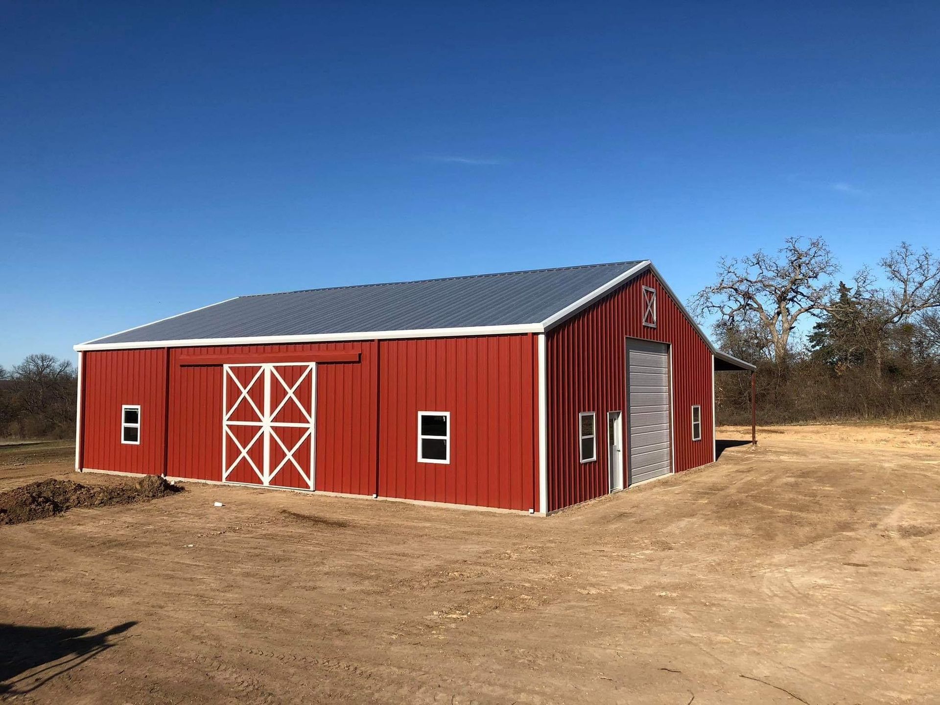 A large red metal barn with a gray roof and white trim, featuring double doors and a roll-up door, set on dirt terrain.