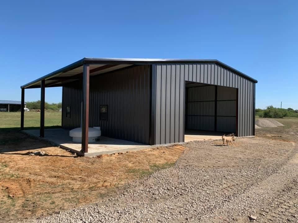 A dark gray metal workshop with a side porch, large open front bay, and a concrete pad on a rural, gravel-covered lot.