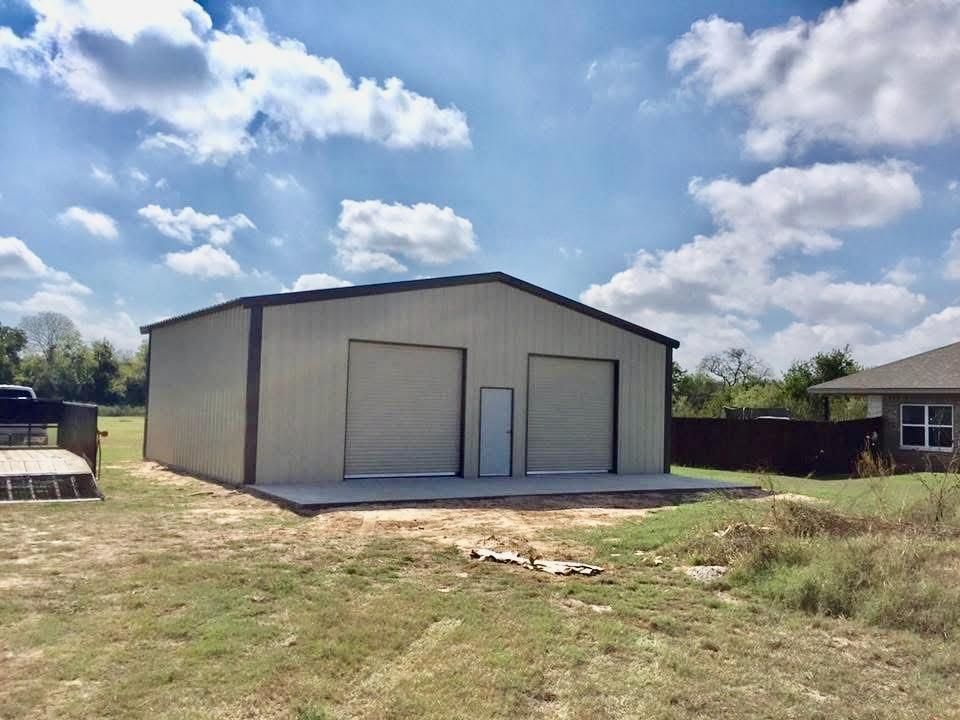 A light-colored metal workshop building with two roll-up doors and a center entrance on a rural lot under a cloudy sky.