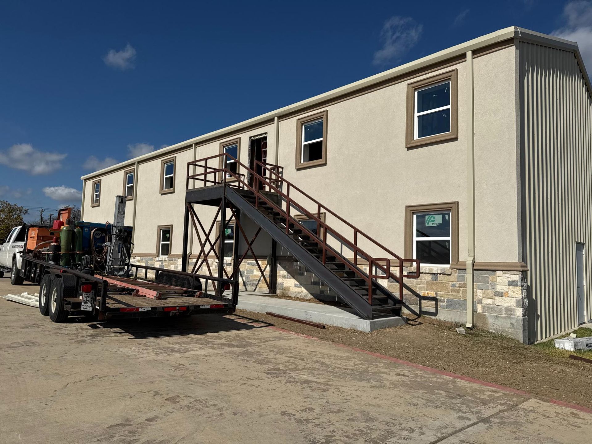 Two-story modular building with tan siding, stone base, and an exterior metal staircase. A utility trailer is parked nearby.