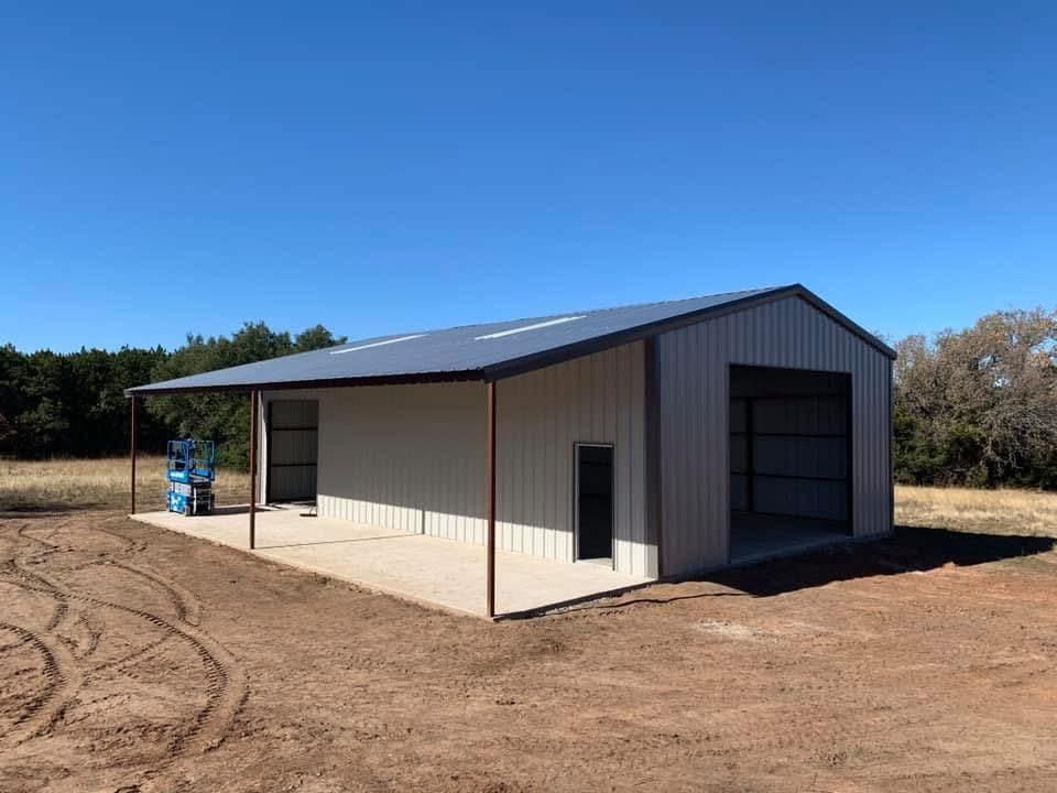 A metal pole barn with light walls, a dark roof, and an open bay, situated in a field under a clear blue sky.
