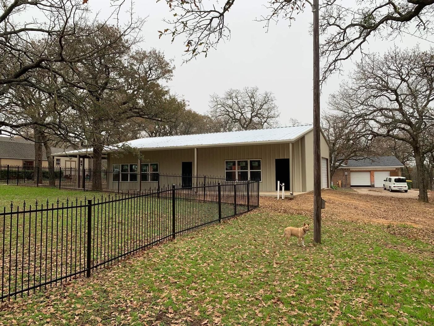 A single-story, light-colored metal house with a metal roof, front porch, and a fenced yard with trees, seen in daylight.