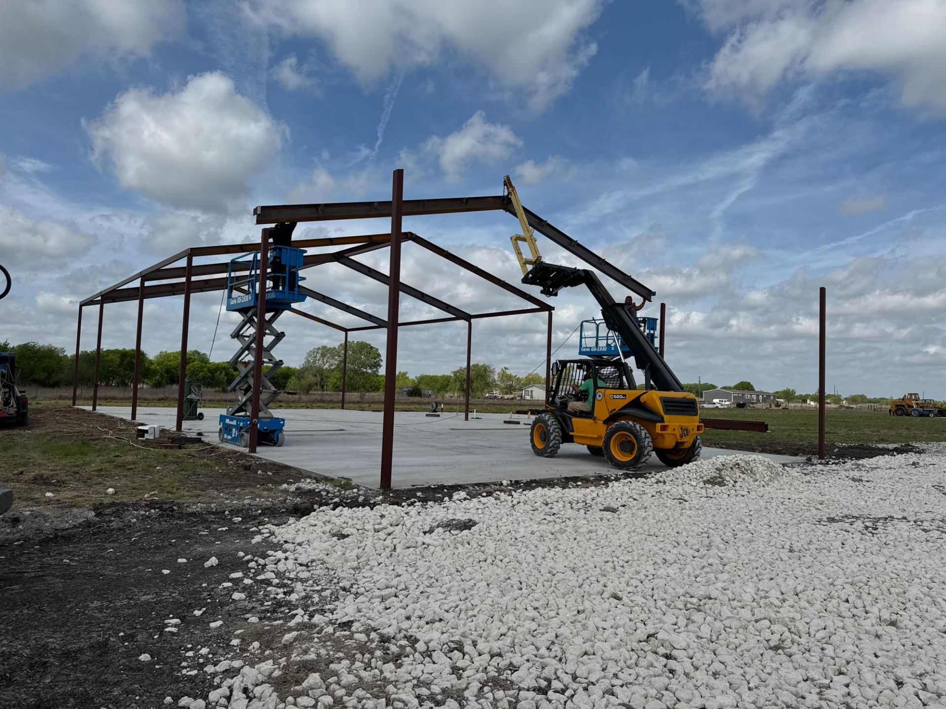 Construction equipment, including a yellow telehandler and a blue scissor lift, erecting the steel frame of a building.