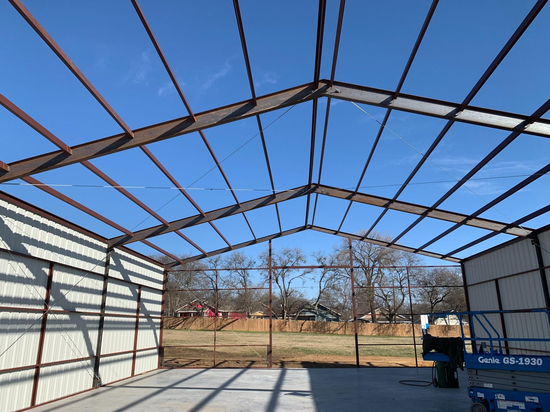 Construction of a metal building frame with exposed steel beams, walls on the sides, and an open front against a blue sky.