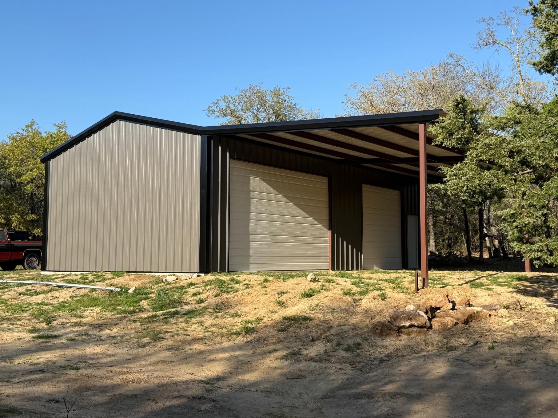 A metal garage with a side carport, featuring light beige vertical siding and two garage doors, set in a wooded area.