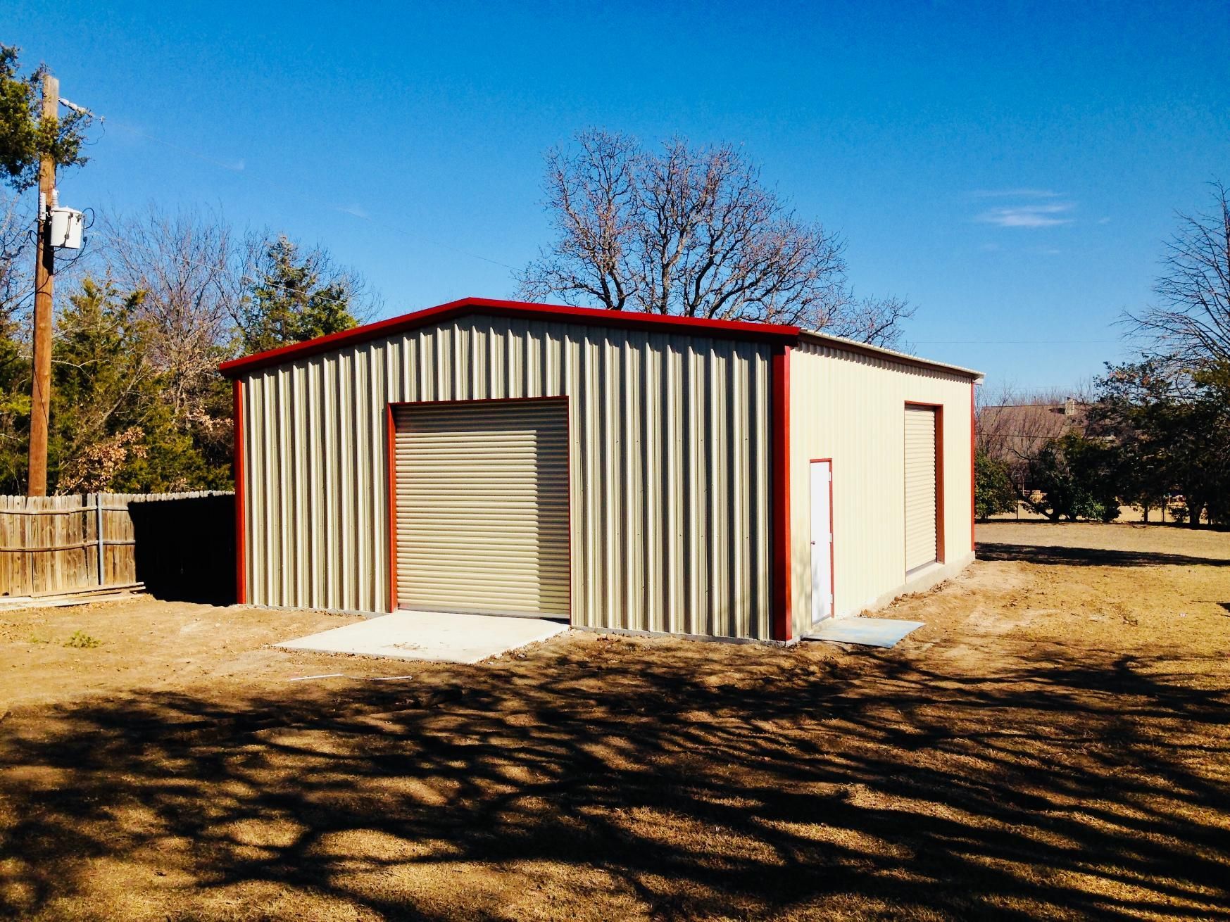 A tan metal garage with red trim, a roll-up door, and a side entry door, situated in a dry, grassy field under a blue sky.