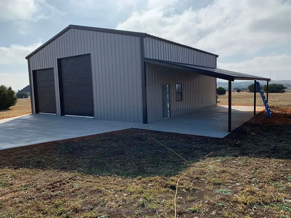 A tan metal workshop with a side lean-to, two black garage doors, a side door, and a concrete pad in a rural setting.