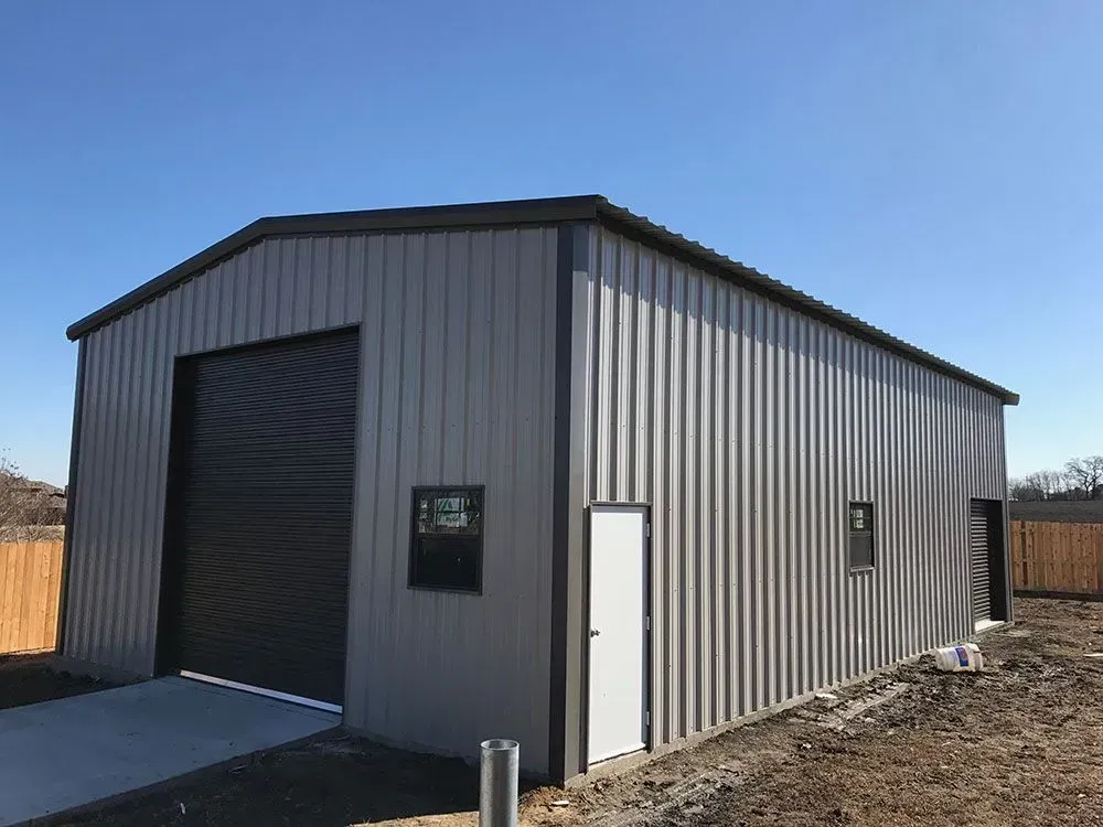 A gray metal building with a dark roll-up door, a white side door, and two windows against a clear blue sky.