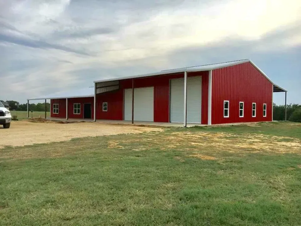 A red metal building with two garage doors and multiple windows, situated on a grassy lot under a cloudy sky.