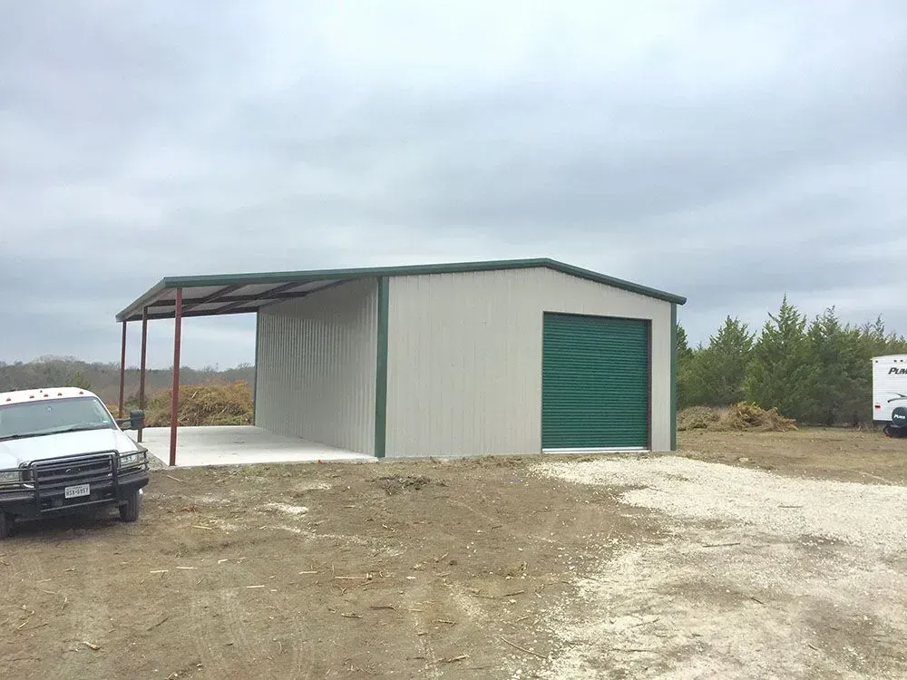 A tan metal building with a green garage door and attached covered carport, situated on a dirt lot under a cloudy sky.