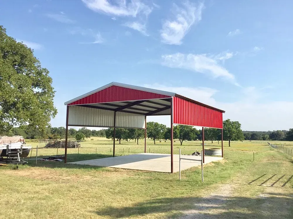 A metal carport with a red roof and partial side walls stands on a concrete slab in a grassy, open field under a blue sky.