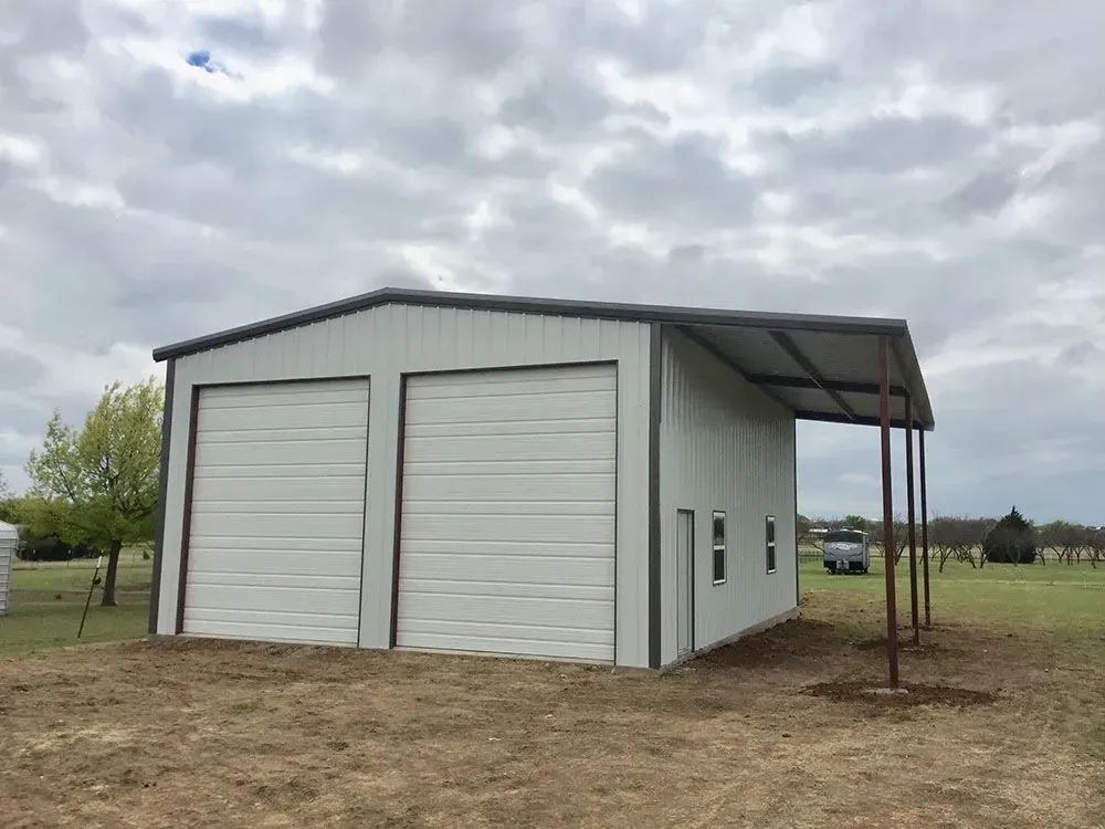 A modern white metal garage with two large roll-up doors and a side lean-to shelter, situated on a grassy field.