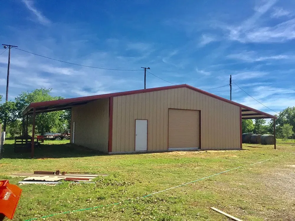 A tan metal barn with red trim, a single entry door, and a large garage door, situated in a grassy field under a blue sky.
