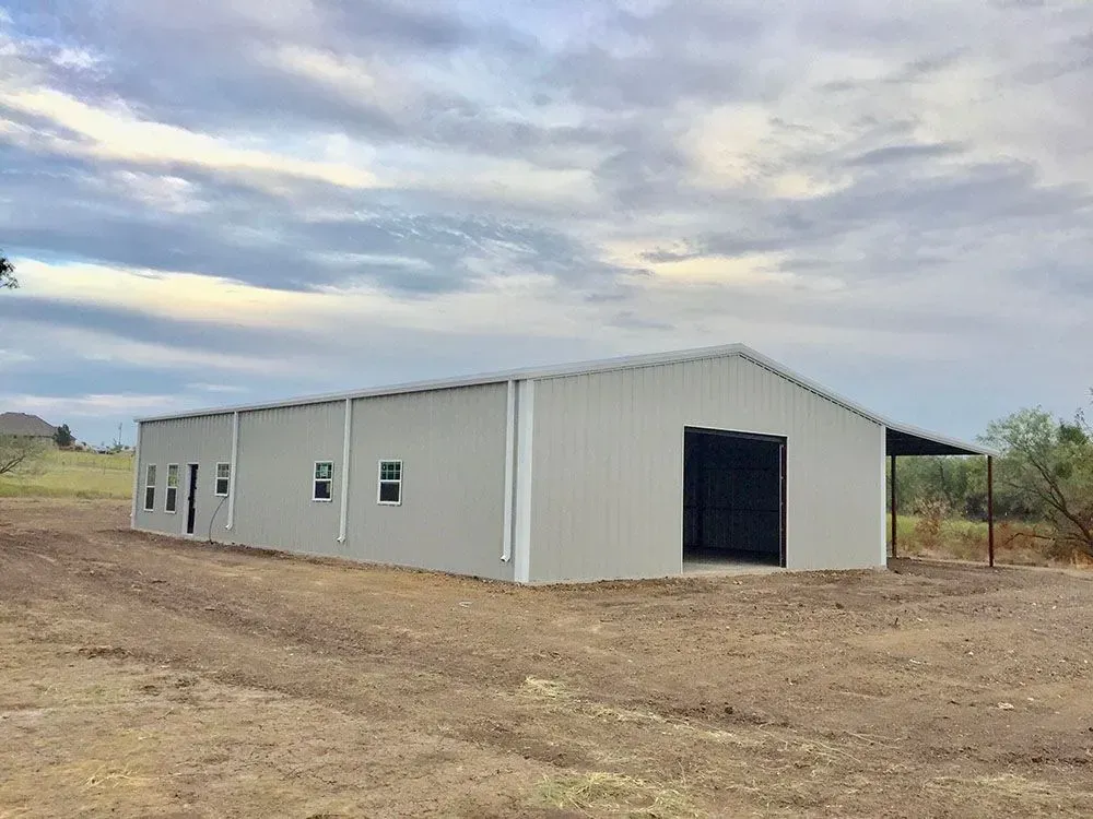 A long, light-gray metal building with several windows and a large open doorway sits on a dirt lot under a cloudy sky.